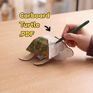 Op de afbeelding: Een kartonnen schildpad knutselproject met een gedeeltelijk ingekleurd schild met groene en bruine geometrische patronen. De tekst 'Carboard Turtle .PDF' is erboven zichtbaar en een groene stift wordt gebruikt om de witte delen in te kleuren.