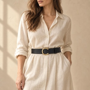 Peut inclure: Une robe chemise beige à manches longues en tissu texturé, cintrée à la taille par une large ceinture en cuir noir dotée d'une boucle métallique dorée arrondie et proéminente qui constitue le point focal central de la tenue.