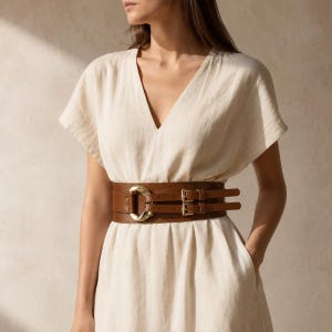 Puede incluir: Un vestido color crema de manga corta con escote en V, combinado con un cinturón ancho de color marrón. El cinturón presenta dos correas horizontales, dos hebillas rectangulares doradas y un gran anillo de metal dorado redondeado en el centro.