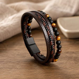 May include: A multi-strand bracelet featuring a combination of dark brown braided leather cords, smooth matte black beads, and polished tiger eye gemstone beads. The bracelet is secured with a rectangular matte black magnetic metal clasp.