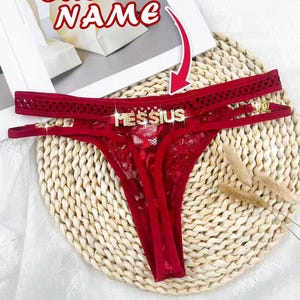Peut inclure: Un string en dentelle rouge foncé orné d'une plaque nominative en métal doré portant l'inscription MESSIUS en lettres capitales. Le vêtement présente des lanières latérales fines et un motif en dentelle texturée sur le devant, avec une flèche rouge.