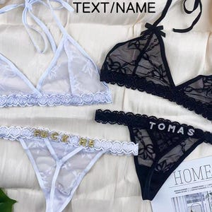 Pode incluir: Um conjunto de bralette e fio dental em renda branca com letras douradas FUCK ME, ao lado de um conjunto em renda preta com letras prateadas TOMAS. Ambos os conjuntos possuem alças estilo frente única e padrões florais delicados.