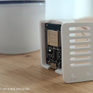 Könnte beinhalten: Ein schwarzes ESP32-Mikrocontroller-Board in einem weißen, 3D-gedruckten Kunststoffgehäuse mit passendem, abnehmbarem Lüftungsdeckel. Der Deckel hat horizontale Schlitze und den geprägten Text EN und BOOT am unteren Rand der Vorderseite.