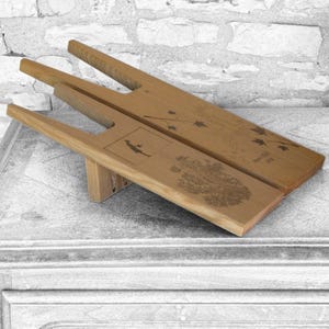 May include: Two light brown wooden boot jacks with a U-shaped cutout at one end. The top boot jack features a dark engraved branch with leaves and the text 'Savoie 2018'. The bottom boot jack displays an engraved silhouette of a person and a tree.