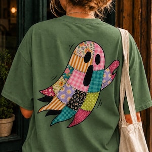 May include: An olive green t-shirt featuring a large graphic on the back of a ghost made of various colorful patchwork patterns, including floral prints, gingham, and polka dots. The ghost has two black oval eyes and an open black mouth.