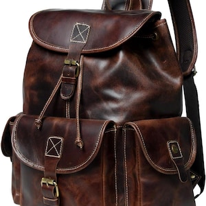May include: A dark brown leather backpack featuring a main compartment with a flap closure, a drawstring, and a buckle strap. The front includes two exterior pockets with flap closures and buckle straps, complemented by white contrast stitching.