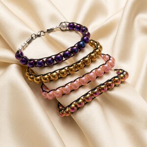 Può includere: Quattro braccialetti con perline rotonde in finiture metalliche viola, oro, rosa chiaro e iridescenti. Ogni braccialetto è realizzato con un cordino nero intrecciato tra le perline e include una chiusura a moschettone in metallo.
