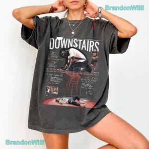 Op de afbeelding: Een antracietgrijs oversized T-shirt met de witte tekst DOWNSTAIRS. De afbeelding bevat een persoon op een trap, een persoon liggend op een rode vloer en handgeschreven notities. Het ontwerp heeft een verweerde, vintage uitstraling.