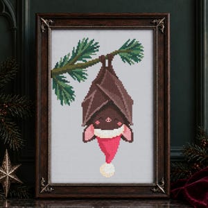 Può includere: Un ricamo a punto croce in una cornice marrone scuro decorata raffigurante un pipistrello marrone appeso a testa in giù a un ramo di pino verde. Il pipistrello indossa un cappello da Babbo Natale rosso con pon pon e bordo bianchi.