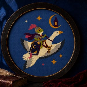 May include: A circular framed cross-stitch embroidery on a dark blue background featuring a green frog knight in armor with a pink plume and purple cape riding a white goose. The goose wears a harness and carries a dark blue book with a gold crescent moon.