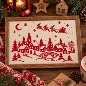 May include: A rectangular cross-stitch embroidery in a wooden frame featuring a red winter village scene on a cream fabric. The design depicts houses, pine trees, a stone bridge, and a crescent moon with Santa in a sleigh.