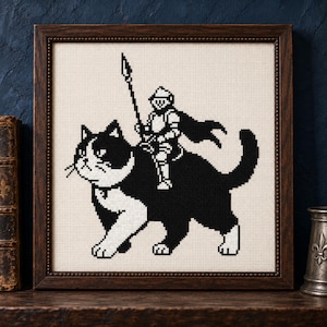 May include: A framed cross-stitch embroidery featuring a black and white cat with a knight in silver armor riding on its back. The knight holds a tall spear and wears a cape. The design is rendered in a pixelated style on a cream-colored fabric.