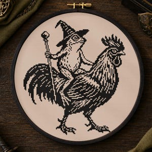 May include: A circular embroidery hoop containing a cross-stitch design on light beige fabric. The black thread pattern depicts a frog wearing a pointed wizard hat and holding a staff while riding on the back of a large standing rooster.