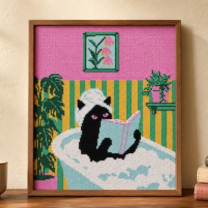 May include: A framed cross-stitch embroidery depicting a black cat with a white towel on its head, reading a light blue book while sitting in a bubble-filled bathtub. The background features yellow and green vertical stripes and a pink wall.