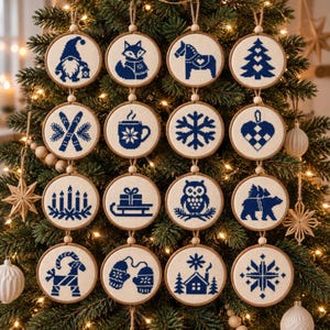 May include: A set of sixteen circular wooden embroidery hoops featuring blue cross-stitch designs on white fabric. The patterns include a gnome, fox, horse, tree, skis, mug, snowflake, heart, candles, sled, owl, bear, ram, mittens, house, and star.