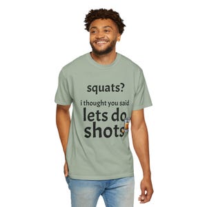 May include: A sage green short-sleeved t-shirt featuring the black text "squats? i thought you said lets do shots" with a small graphic of a shot glass next to the word shots. The shirt has a crew neckline and a standard fit.
