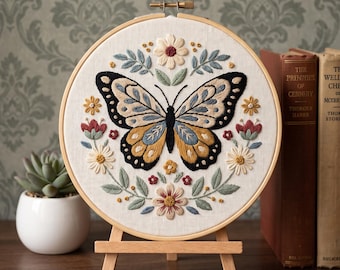 May include: A circular wooden embroidery hoop containing a fabric panel with a hand-stitched butterfly design in shades of black, yellow, and blue, surrounded by a symmetrical arrangement of embroidered flowers and leaves in muted earth tones.