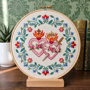 May include: A circular wooden embroidery hoop containing white fabric with a hand-stitched design of two hearts. One heart features a cross and flames, while the other displays a crown, flames, and a dagger. A floral wreath border surrounds them.