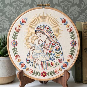 Op de afbeelding: Een ronde houten borduurring met een stoffen paneel met een handgestikt ontwerp van een persoon die een kind vasthoudt. De figuren zijn omringd door een gouden halo en een bloemenkrans met rode, blauwe en groene draadaccenten.