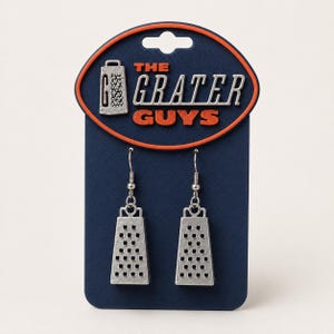 May include: A pair of silver-toned metal cheese grater earrings attached to a dark blue display card. The card features the text THE GRATER GUYS in silver and orange lettering, along with a small silver grater icon at the top left corner.