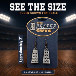 May include: A pair of silver-colored cheese grater earrings on a dark blue display card labeled The Grater Guys. The earrings feature a textured metal design with hook-style backings. The card indicates the items are approximately 2 inches in size.