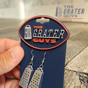 May include: A pair of silver-colored cheese grater earrings attached to a dark blue display card. The card features an orange-bordered oval logo with the text THE GRATER GUYS and a small graphic of a grater. The earrings hang from metal hooks.