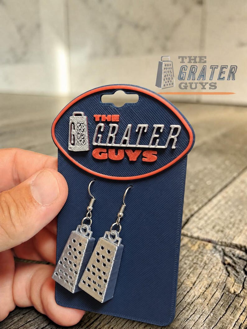 May include: A pair of silver-colored cheese grater earrings attached to a dark blue display card. The card features an orange-bordered oval logo with the text THE GRATER GUYS and a small graphic of a grater. The earrings hang from metal hooks.