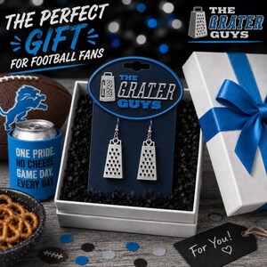 May include: A pair of silver-toned cheese grater earrings displayed on a dark blue branded card labeled The Grater Guys, presented in a white gift box with a blue ribbon, alongside a football, a blue can cooler, and a bowl of pretzels.