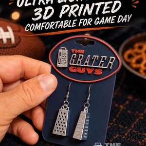 May include: A pair of silver-colored, 3D printed cheese grater earrings attached to a dark blue display card. The card features the text 'THE GRATER GUYS' in red and white lettering. The earrings hang from silver-toned metal hooks.