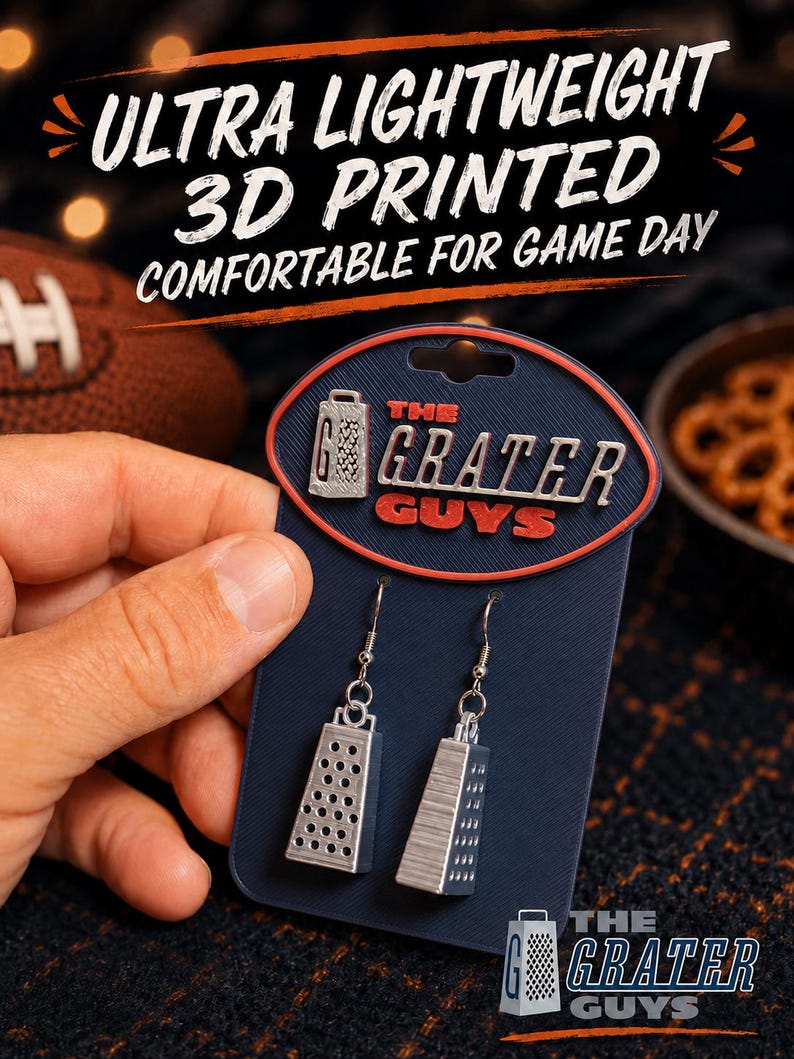 May include: A pair of silver-colored, 3D printed cheese grater earrings attached to a dark blue display card. The card features the text 'THE GRATER GUYS' in red and white lettering. The earrings hang from silver-toned metal hooks.