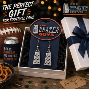 May include: A pair of silver-toned cheese grater earrings attached to a dark blue display card with the text THE GRATER GUYS. The earrings feature a classic four-sided grater shape with a textured surface and are suspended from silver-toned hooks.