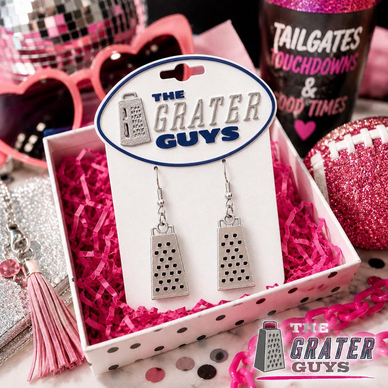 May include: Silver-toned cheese grater dangle earrings attached to a white display card featuring the text THE GRATER GUYS. The earrings are presented in a white box filled with pink shredded paper, surrounded by pink accessories and decor.