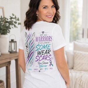 May include: A white t-shirt featuring a graphic on the back with a zebra head illustration in purple and teal, accompanied by the text "Not all warriors wear armor, some wear scars" and "Rare disease warrior" with various medical acronyms.