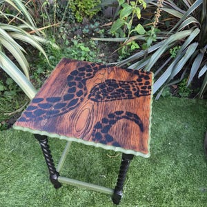 May include: A square wooden side table featuring a natural wood grain top with a black painted octopus tentacle design. The table has four legs, with two featuring a twisted spiral texture painted black and two straight legs painted light green.