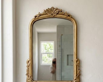 May include: An arched wall mirror featuring a gold-toned frame with ornate, carved scrollwork detailing at the top and bottom. The mirror has a smooth, reflective glass surface that displays a clear view of the surrounding interior space.