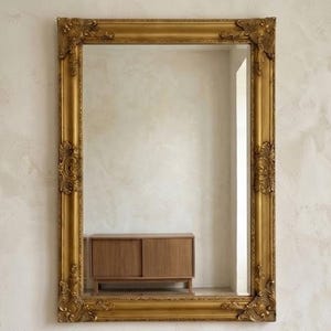 Pode incluir: Um espelho de parede retangular com uma moldura dourada larga e ornamentada, apresentando detalhes esculpidos intrincados em cada canto e ao longo das laterais. A superfície de vidro reflete um armário de madeira com portas de correr.