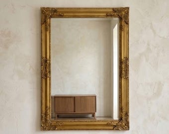 Peut inclure: Un miroir mural rectangulaire doté d'un large cadre doré orné de détails sculptés complexes à chaque coin et sur les côtés. La surface en verre réfléchissant offre une vue claire d'une armoire en bois avec des portes coulissantes.
