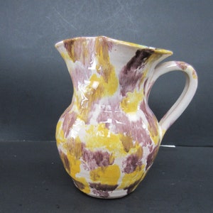 May include: A ceramic pitcher with a wide body and a handle. The pitcher is painted with a pattern of yellow, white, and maroon splotches. The rim of the pitcher has a wavy design.