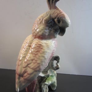 May include: A ceramic parrot figurine in shades of pink, cream, and gray, perched on a branch with green leaves. The bird has a detailed feather design and a curved beak. The figurine is a decorative item.