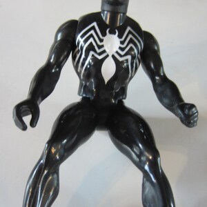 May include: A black action figure of a superhero with a white spider emblem on the chest. The figure has white eye details and a white spiderweb design on the chest and back. The figure is posed with legs spread and arms out.