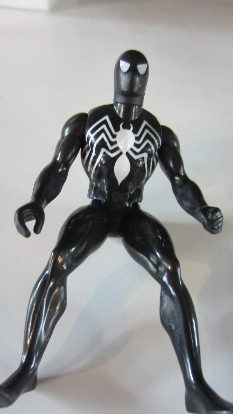 May include: A black action figure of a superhero with a white spider emblem on the chest. The figure has white eye details and a white spiderweb design on the chest and back. The figure is posed with legs spread and arms out.