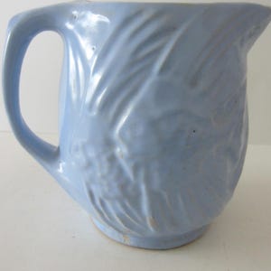 May include: A light blue ceramic pitcher with a curved handle and a spout. The pitcher has a textured, embossed design on the body. The pitcher is likely used for serving beverages.