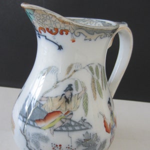 May include: White porcelain pitcher with a curved handle and spout. The pitcher is decorated with a hand-painted scene featuring figures in traditional clothing, foliage, and architectural elements. The rim has a decorative border.