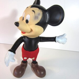 Pode incluir: Uma figura vintage do Mickey Mouse. A figura tem um corpo, cabe&ccedil;a e orelhas pretas, com um rosto bronzeado, cal&ccedil;&otilde;es vermelhos e sapatos castanhos. Luvas brancas nas m&atilde;os. A figura est&aacute; posando com um bra&ccedil;o estendido.