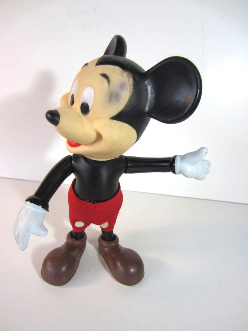 Pode incluir: Uma figura vintage do Mickey Mouse. A figura tem um corpo, cabe&ccedil;a e orelhas pretas, com um rosto bronzeado, cal&ccedil;&otilde;es vermelhos e sapatos castanhos. Luvas brancas nas m&atilde;os. A figura est&aacute; posando com um bra&ccedil;o estendido.