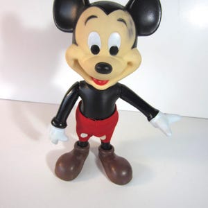 Pode incluir: Uma figura vintage do Mickey Mouse. A figura tem um corpo, orelhas e bra&ccedil;os pretos, com um rosto bronzeado, luvas brancas e sapatos castanhos. Mickey usa cal&ccedil;&otilde;es vermelhos. A figura est&aacute; posada com os bra&ccedil;os estendidos.