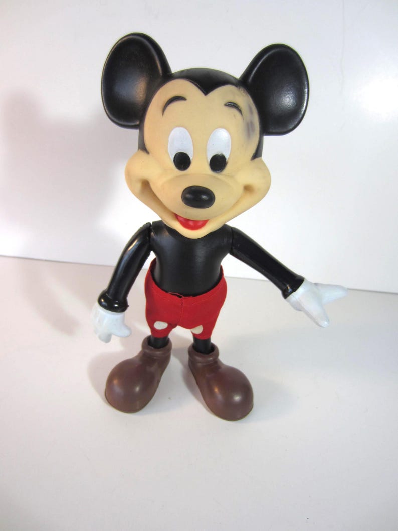 Pode incluir: Uma figura vintage do Mickey Mouse. A figura tem um corpo, orelhas e bra&ccedil;os pretos, com um rosto bronzeado, luvas brancas e sapatos castanhos. Mickey usa cal&ccedil;&otilde;es vermelhos. A figura est&aacute; posada com os bra&ccedil;os estendidos.