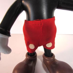 Pode incluir: Uma figura vintage do Mickey Mouse, com corpo, bra&ccedil;os e pernas pretas, luvas brancas e sapatos castanhos grandes. A figura usa cal&ccedil;&otilde;es vermelhos com dois bot&otilde;es brancos. Um item colecion&aacute;vel.