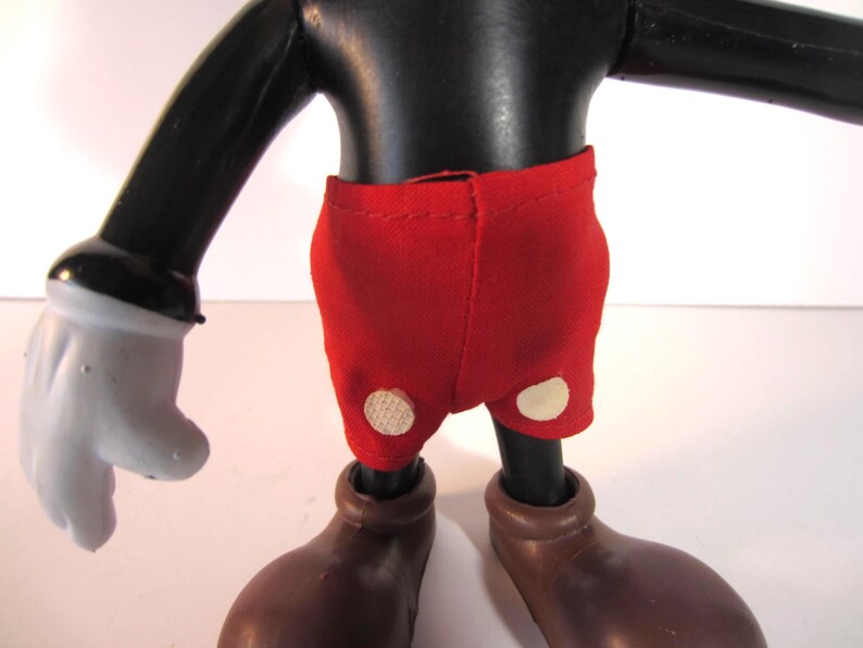 Pode incluir: Uma figura vintage do Mickey Mouse, com corpo, bra&ccedil;os e pernas pretas, luvas brancas e sapatos castanhos grandes. A figura usa cal&ccedil;&otilde;es vermelhos com dois bot&otilde;es brancos. Um item colecion&aacute;vel.