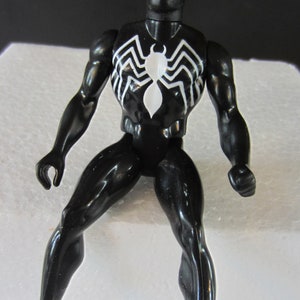 May include: A black action figure of a superhero with a white spider emblem on the chest. The figure has white eye shapes and is posed with one leg extended. The figure is glossy and appears to be made of plastic.